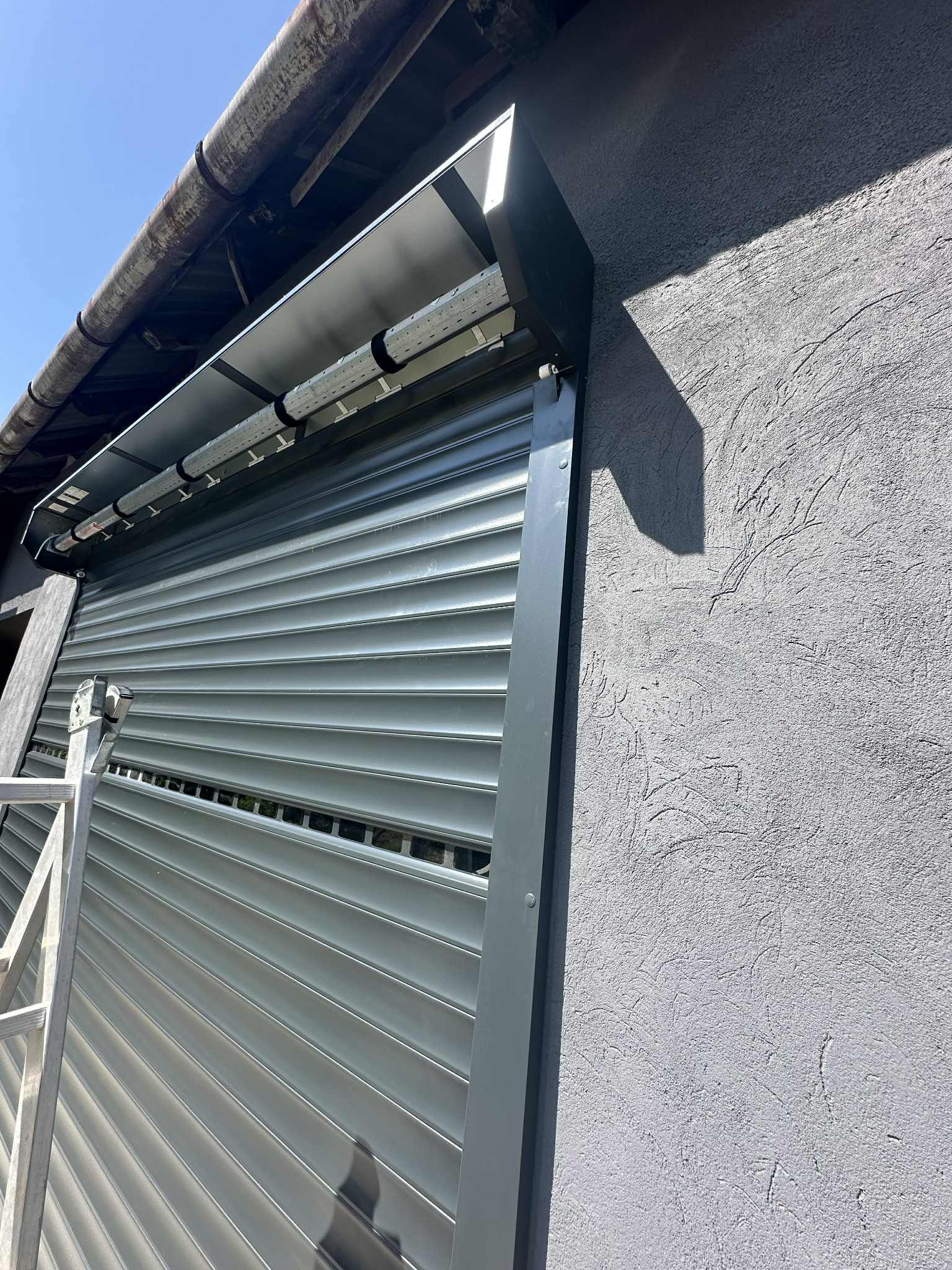 Szara roleta zewnętrzna zamontowana na budynku z szarą, teksturowaną elewacją, widziana z dołu pod kątem. Fragment drabiny po lewej stronie.