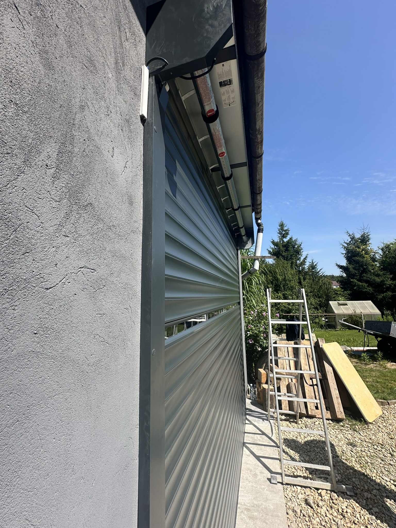 Szara brama rolowana garażowa z widocznym mechanizmem, zamontowana na elewacji budynku. Obok drabina i materiały budowlane. Ujęcie z boku, w tle zieleń.
