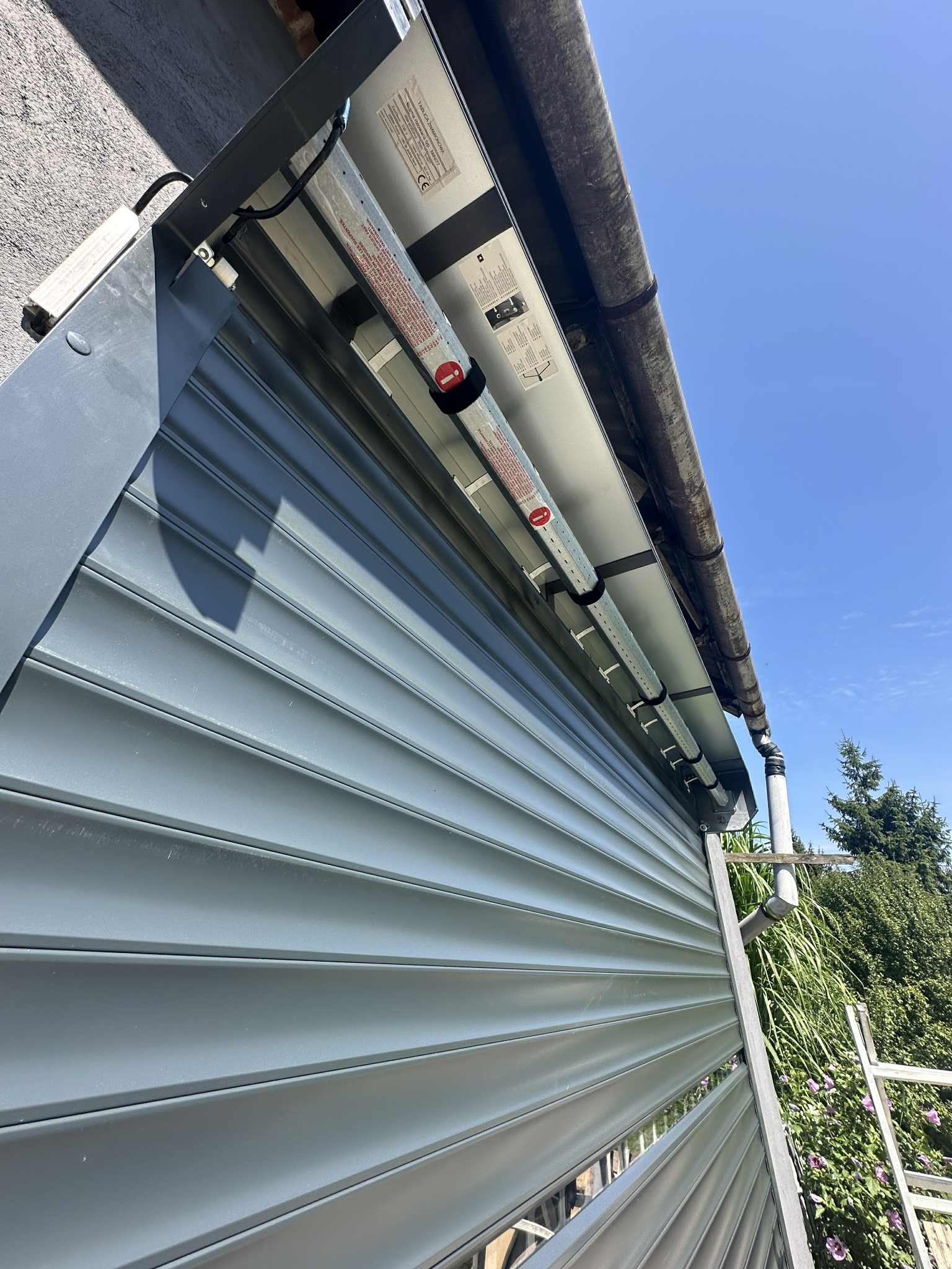 Szara brama garażowa segmentowa z widocznym mechanizmem i elementami montażowymi, sfotografowana pod kątem z dołu, na tle błękitnego nieba i zieleni.