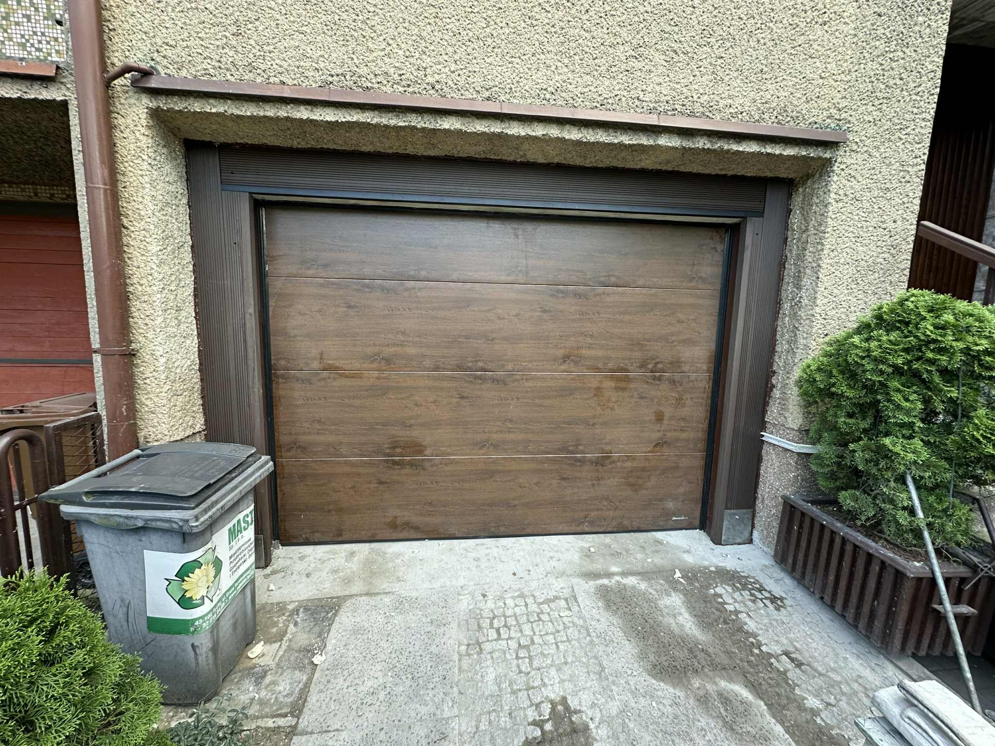 Ciemnobrązowa brama garażowa segmentowa z poziomym wzorem drewna, zamontowana w elewacji budynku. Obok kosz na śmieci i krzew w donicy.
