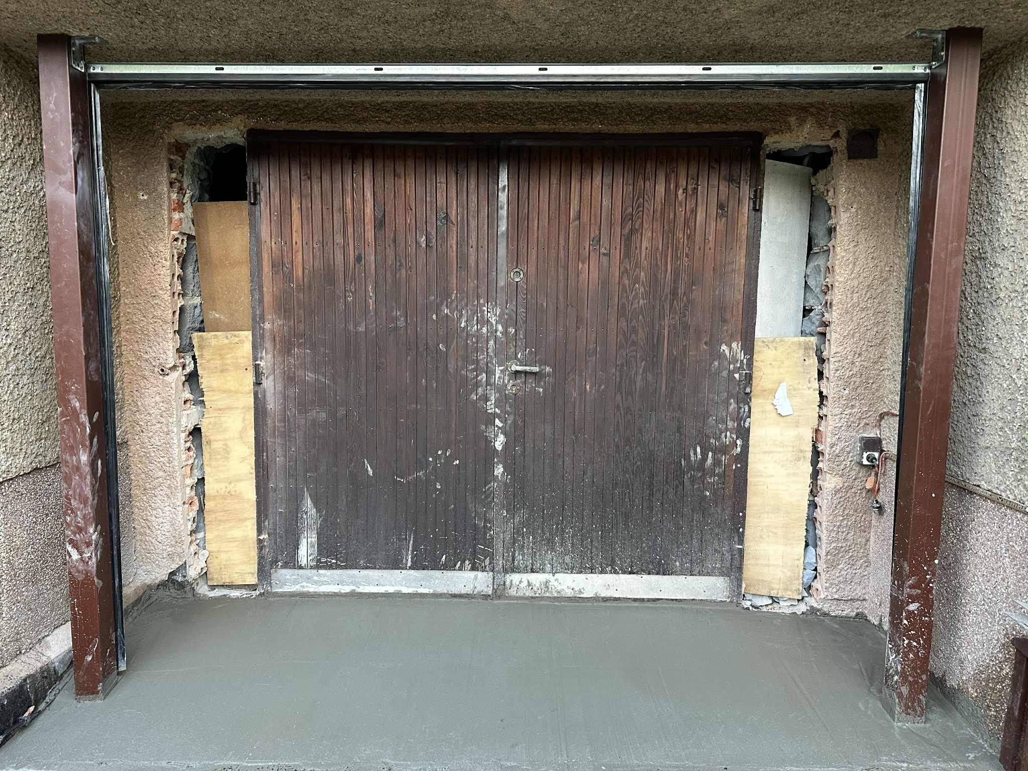 Montaż prowadnic bramy garażowej z drewnianymi drzwiami. Widoczne uszkodzenia muru i świeża wylewka betonowa. Ujęcie z bliska, ukazujące etap prac.