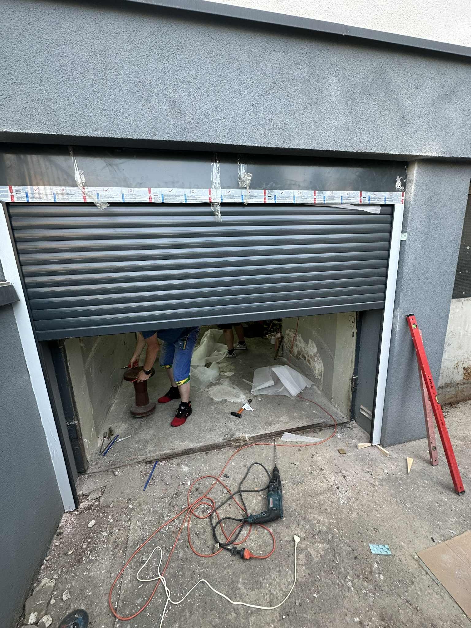 Montaż szarej bramy garażowej rolowanej. Widoczne narzędzia, poziomica i częściowo opuszczona brama. Prace montażowe w trakcie realizacji.