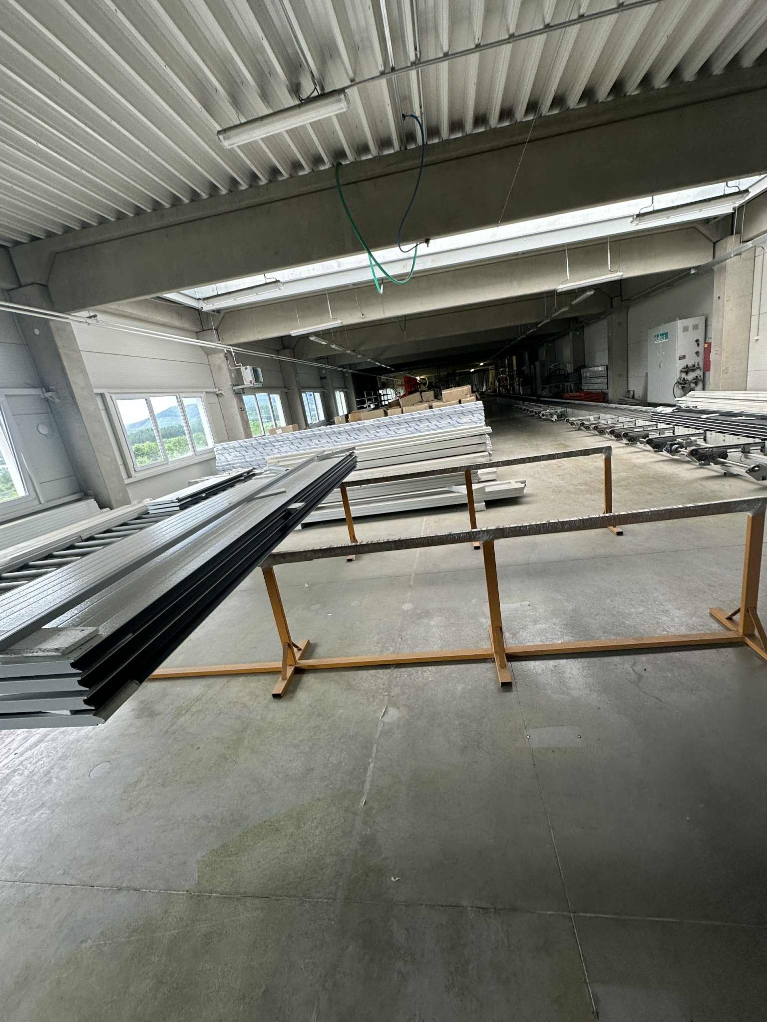 Produkcja okien aluminiowych: stos profili okiennych na metalowym stojaku w hali produkcyjnej z oknami w tle. Widok na linię produkcyjną i sufit hali.