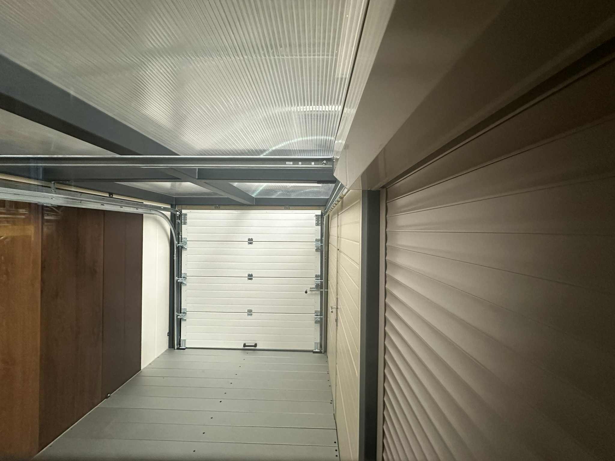 Otwarta, segmentowa brama garażowa wewnątrz garażu z szarą podłogą i metalową konstrukcją sufitu. Widoczne panele boczne i roleta zewnętrzna.