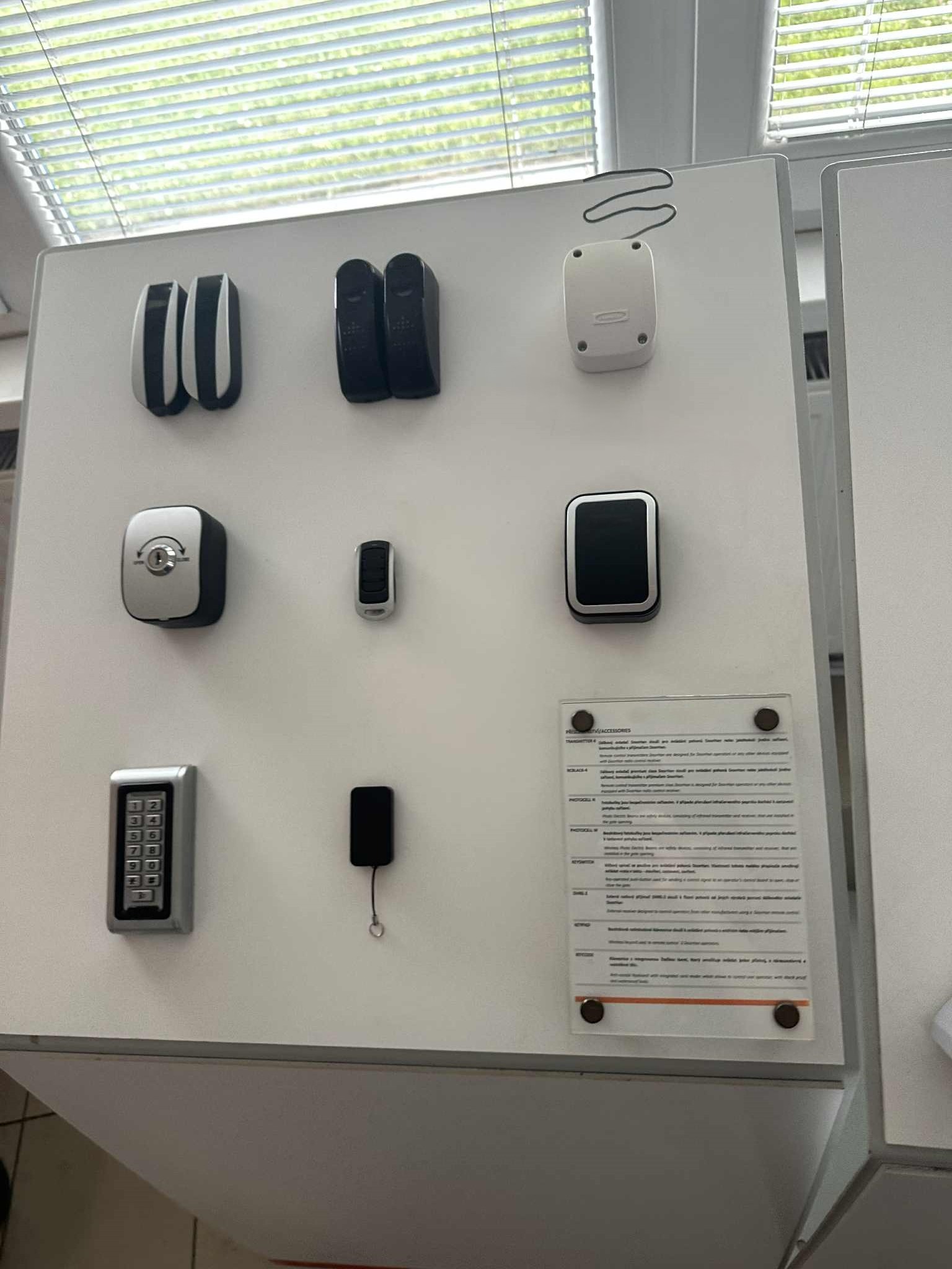 Ekspozycja różnorodnych akcesoriów do bram: piloty, czytniki kart, klawiatury kodowe. Białe tło, okno w tle. Prezentacja asortymentu.