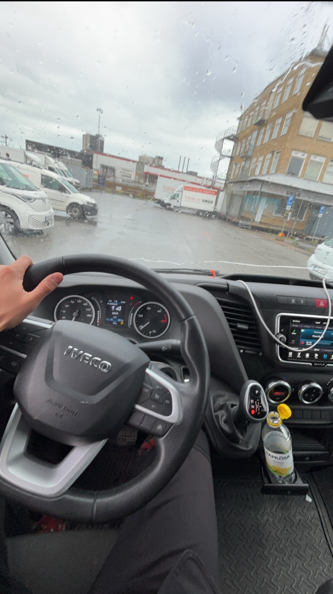 Widok z kabiny Iveco na mokry plac z zaparkowanymi pojazdami. Kierowca trzyma kierownicę, widoczna deska rozdzielcza i butelka wody. Pochmurna pogoda.