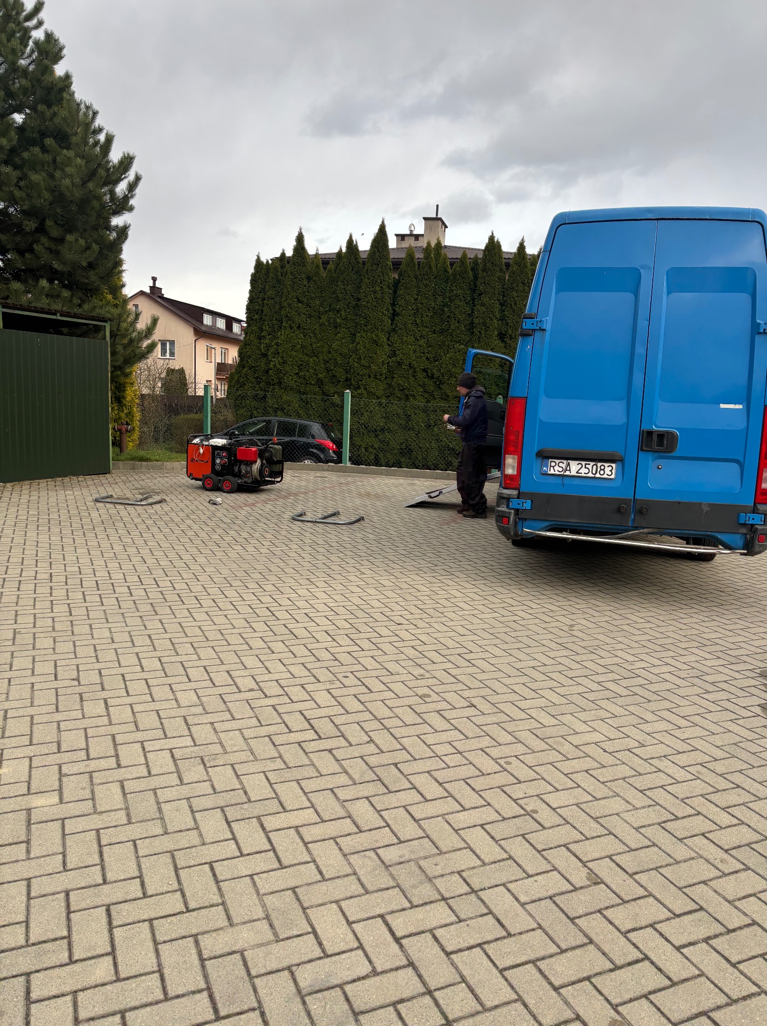 Niebieski bus z otwartymi drzwiami na brukowanym podjeździe, obok agregat prądotwórczy. Osoba wewnątrz busa. Tło: zielony żywopłot i budynek mieszkalny.