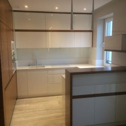 ARCHITEKTURA MEBLI SPÓŁKA Z OGRANICZONĄ ODPOWIEDZIALNOŚCIĄ - Nowoczesna kuchnia z białymi frontami i drewnianymi akcentami, oświetlona lampami wiszącymi. Widoczny blat roboczy i zabudowa kuchenna.