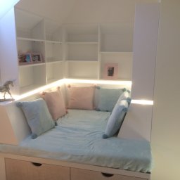 ARCHITEKTURA MEBLI SPÓŁKA Z OGRANICZONĄ ODPOWIEDZIALNOŚCIĄ - Biała zabudowa wnęki na poddaszu z półkami i siedziskiem z szufladami, podświetlona listwą LED, z poduszkami w pastelowych kolorach. Dekoracje w postaci ramek i figurki.