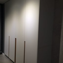ARCHITEKTURA MEBLI SPÓŁKA Z OGRANICZONĄ ODPOWIEDZIALNOŚCIĄ - Długa, biała szafa wnękowa z minimalistycznymi, drewnianymi uchwytami, wkomponowana w szarą ścianę. Oświetlenie górne podkreśla gładkie fronty.