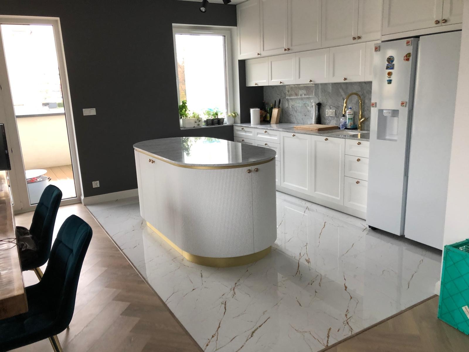 Biała kuchnia z wyspą i marmurowym blatem, złote akcenty, jasne płytki podłogowe z żyłkami, okno z roślinami, nowoczesny design.