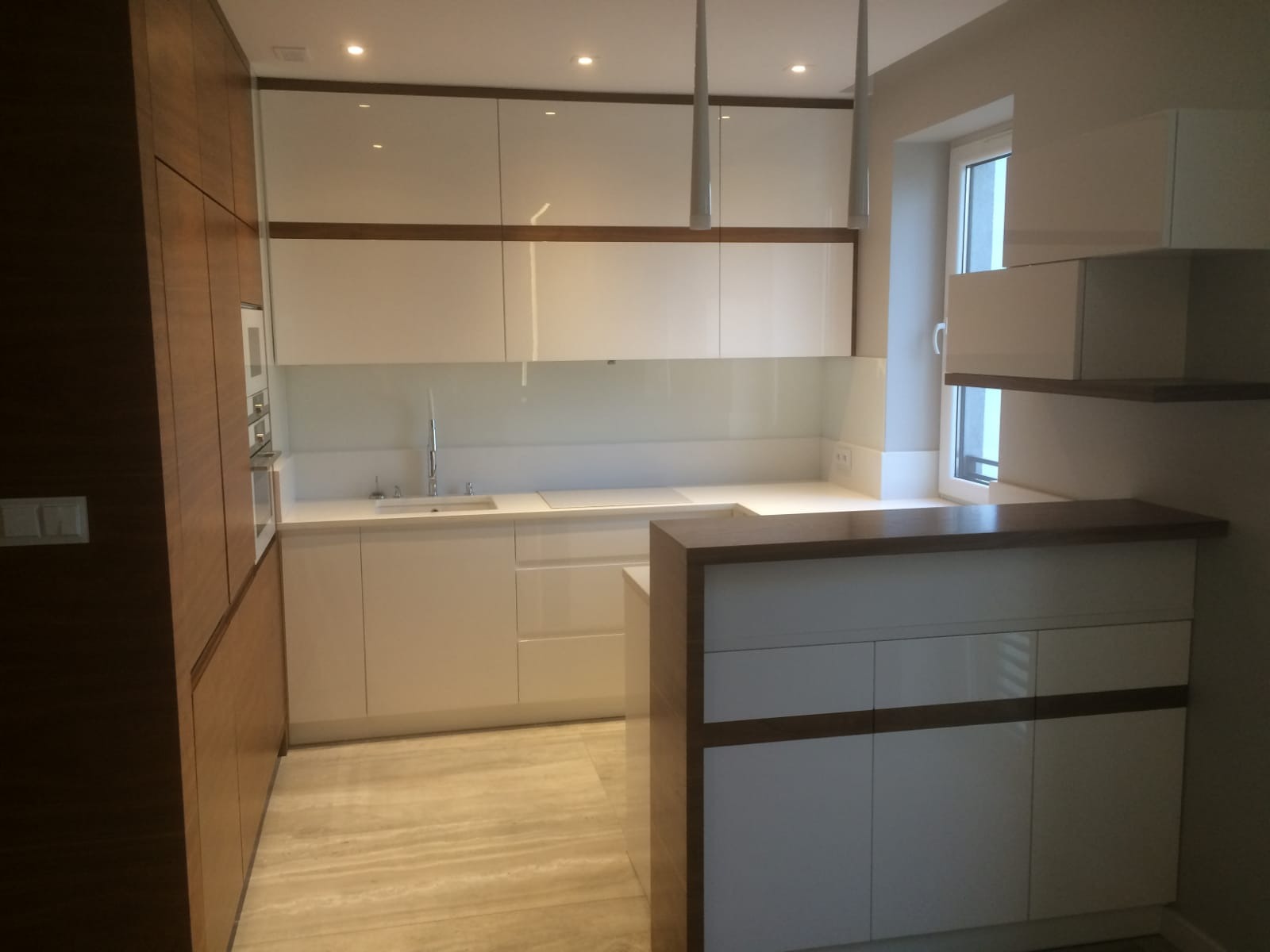 Nowoczesna kuchnia z białymi frontami i drewnianymi akcentami, oświetlona lampami wiszącymi. Widoczny blat roboczy i zabudowa kuchenna.