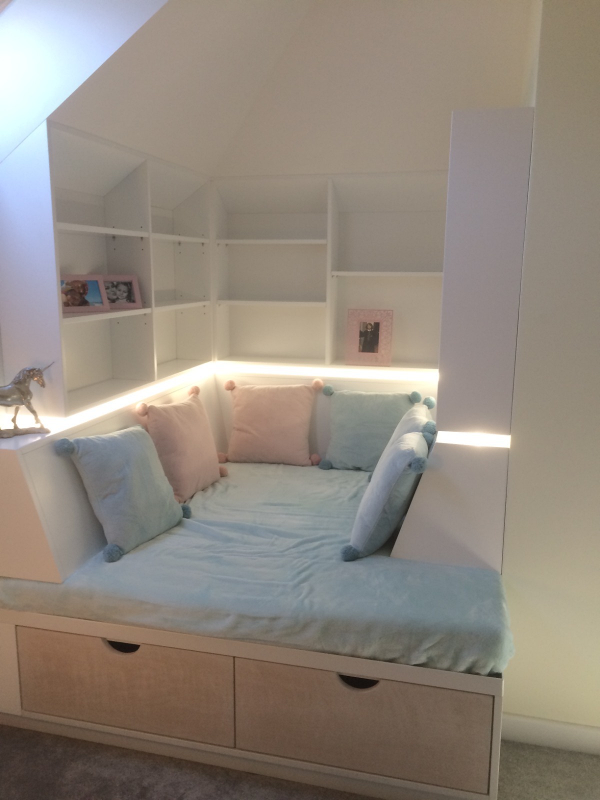 Biała zabudowa wnęki na poddaszu z półkami i siedziskiem z szufladami, podświetlona listwą LED, z poduszkami w pastelowych kolorach. Dekoracje w postaci ramek i figurki.