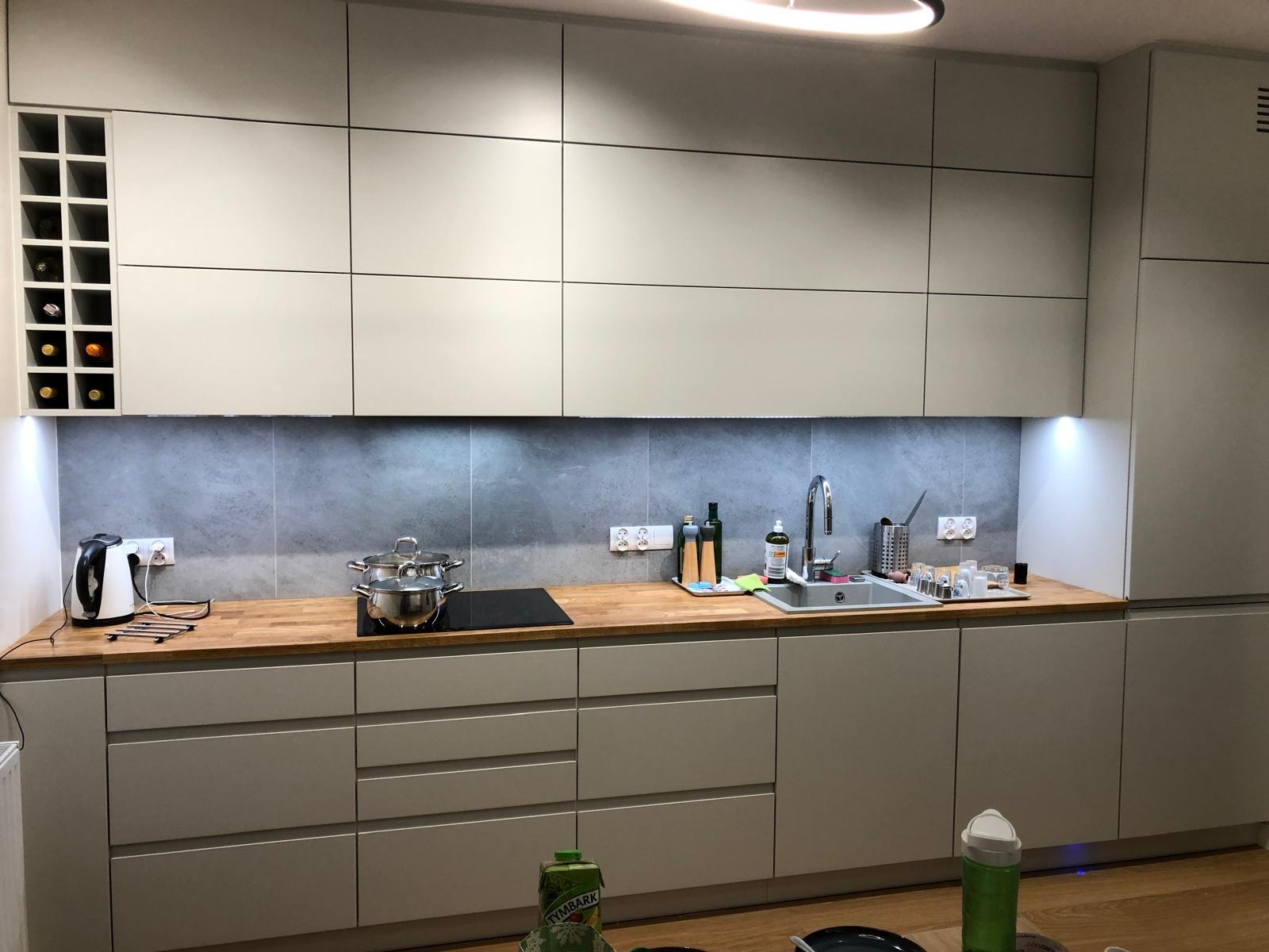 Nowoczesna kuchnia z szarymi frontami, drewnianym blatem i oświetleniem LED. Na blacie czajnik, płyta indukcyjna z garnkami i akcesoria kuchenne. Widoczna zabudowa z szafkami.