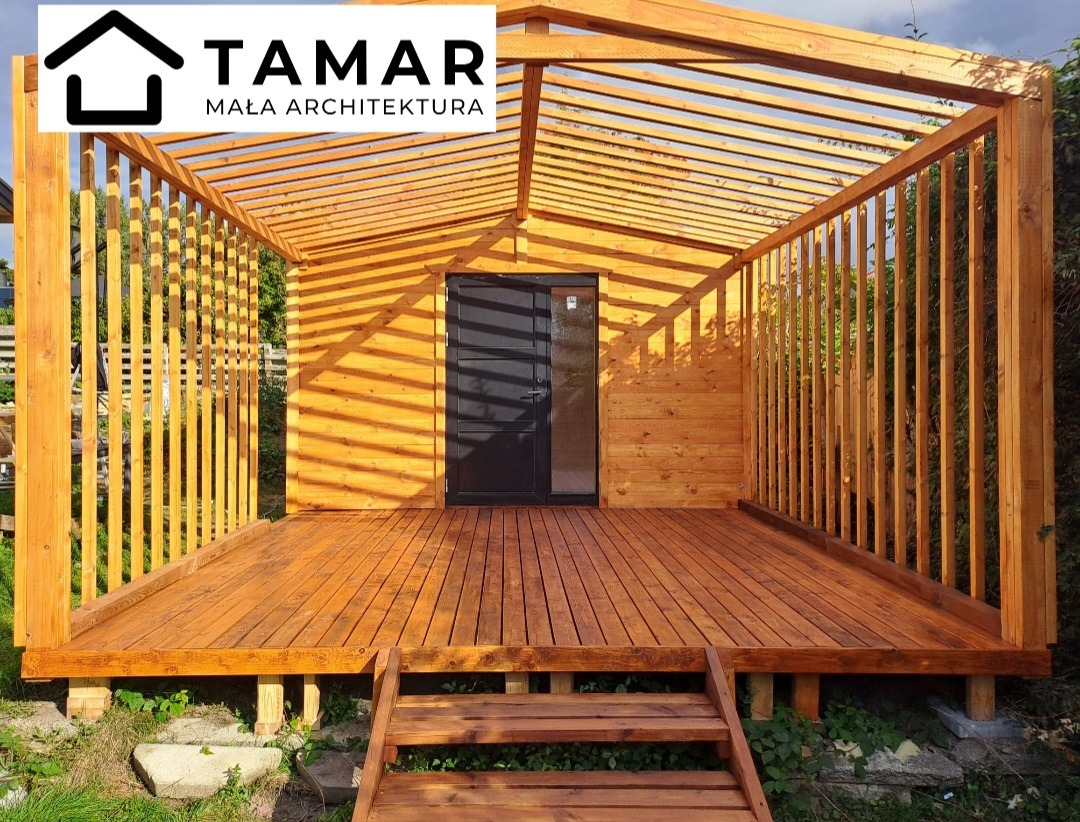 Drewniana altana z zadaszeniem lamelowym i podestem, pomalowana na ciepły brąz, z czarnymi drzwiami wejściowymi. Widoczne schody wejściowe i zieleń w tle.