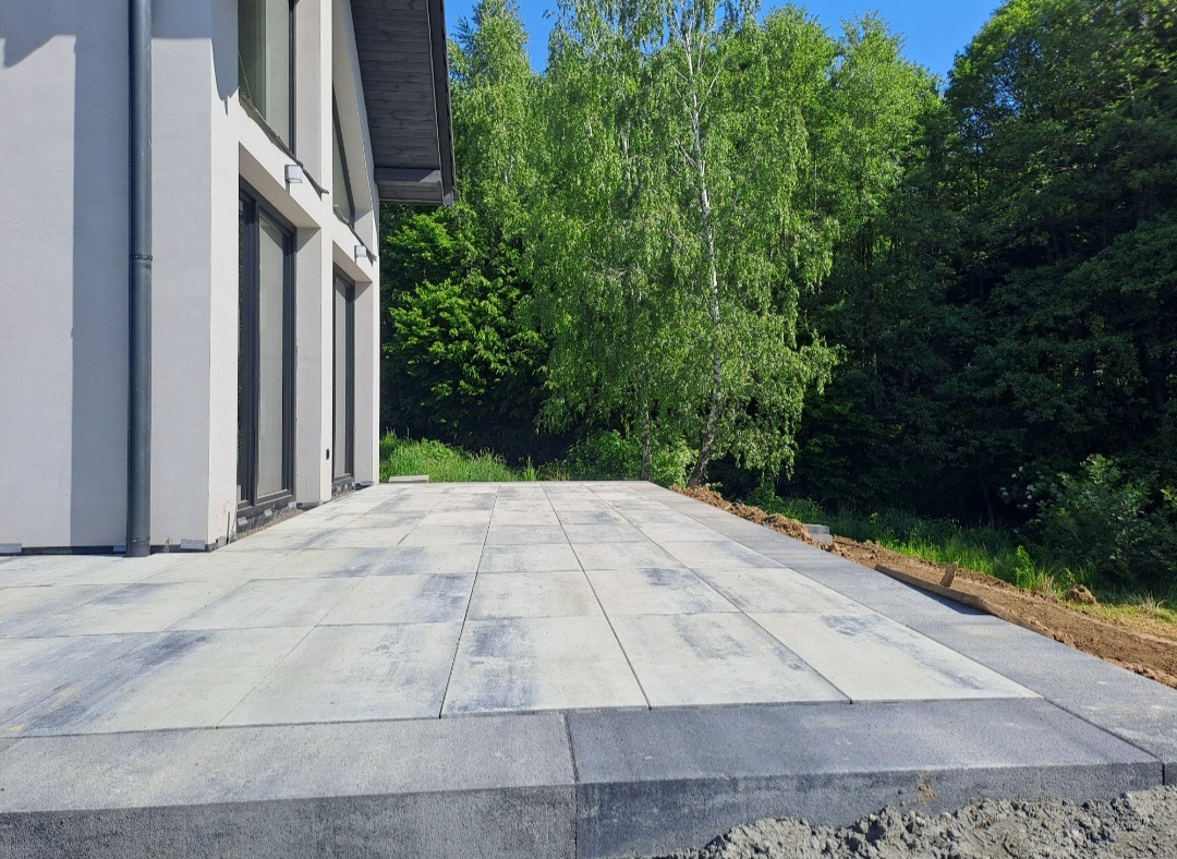 Nowoczesny taras z szarych płyt betonowych, przylegający do białego budynku z dużymi oknami, otoczony zielenią. Widok z niskiej perspektywy.