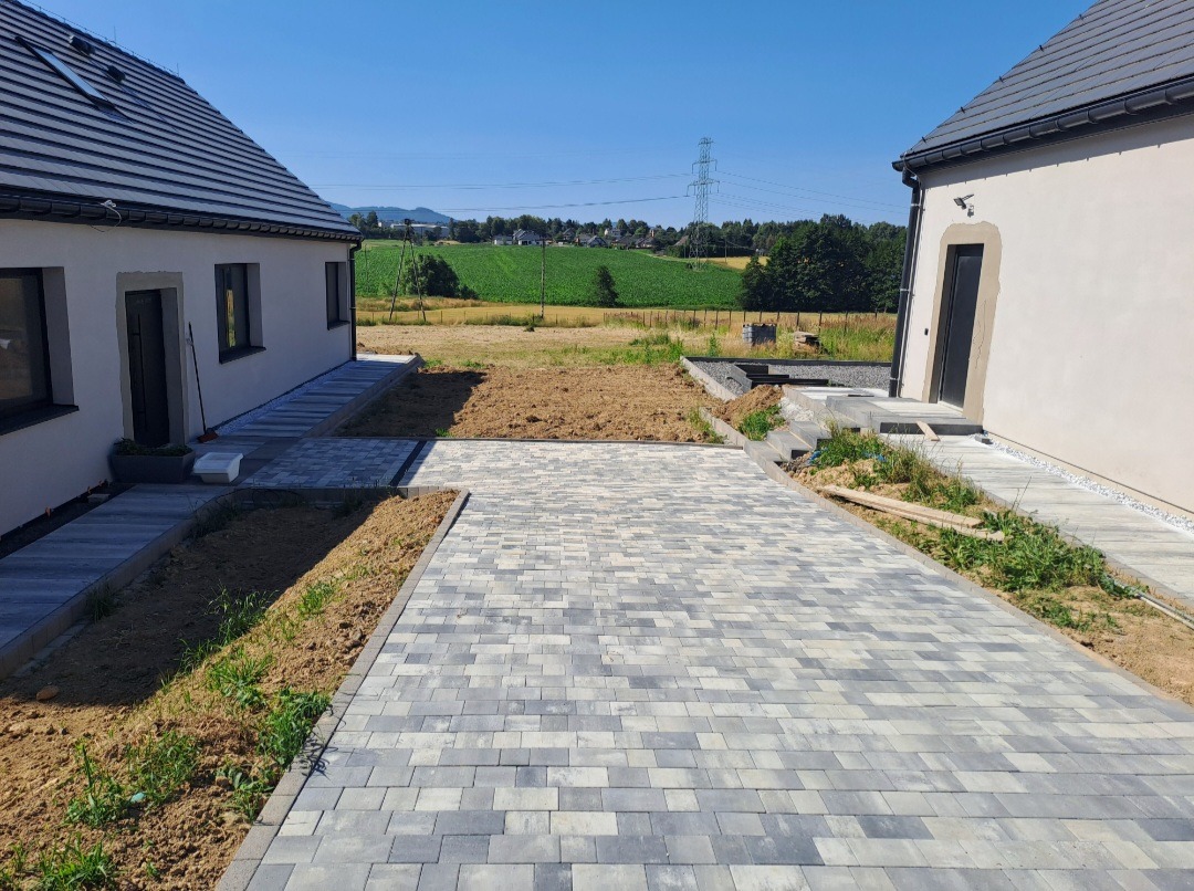 Ułożona szara kostka brukowa na podjeździe między dwoma domami jednorodzinnymi, widok na pole i linię wysokiego napięcia w tle, słoneczny dzień.