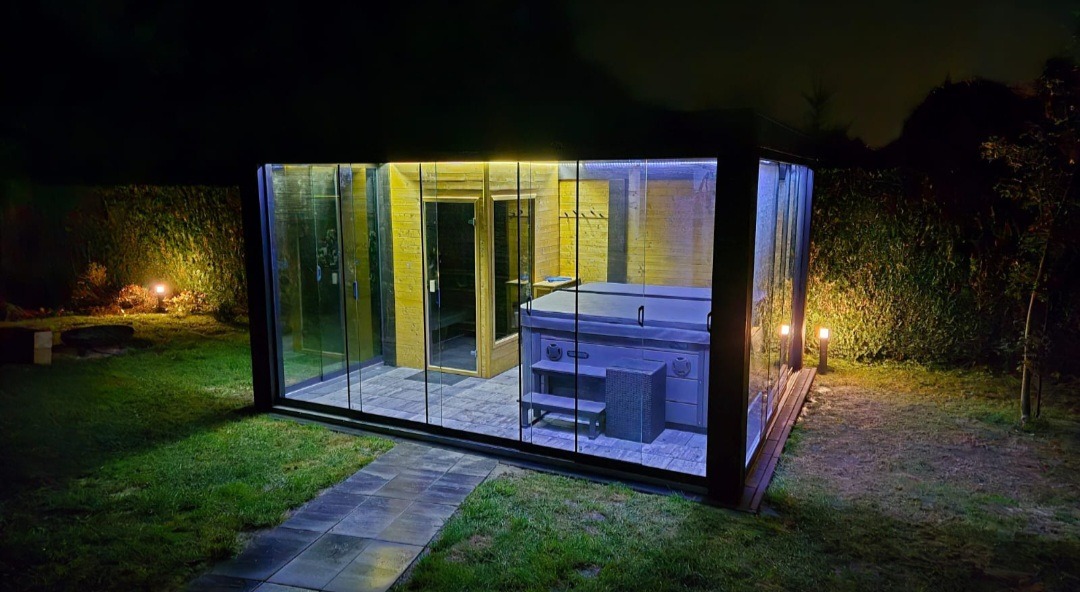 Nowoczesna, przeszklona altana z sauną i jacuzzi, oświetlona dekoracyjnym światłem, otoczona zielenią w nocnej scenerii. Widok z zewnątrz, z bliska.