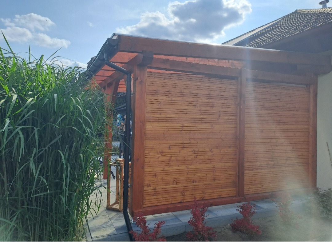 Drewniana pergola z żaluzjowymi panelami, widok z boku, otoczona zielenią. Elementy konstrukcyjne w ciepłym odcieniu brązu, tło z błękitnym niebem i fragmentem dachu.