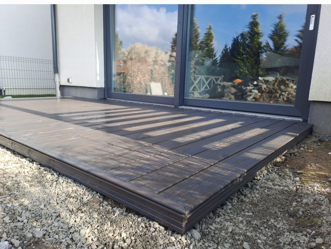 Nowoczesny taras z desek kompozytowych w odcieniach brązu, przylegający do domu z dużym oknem przesuwnym. Widoczne kamienie ozdobne wokół tarasu.