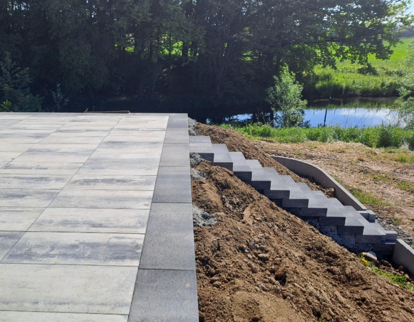 Wykończony taras z szarych płyt i schody z kostki brukowej na zboczu, z widokiem na rzekę i zieleń w tle. Nowoczesny design i staranne wykonanie.