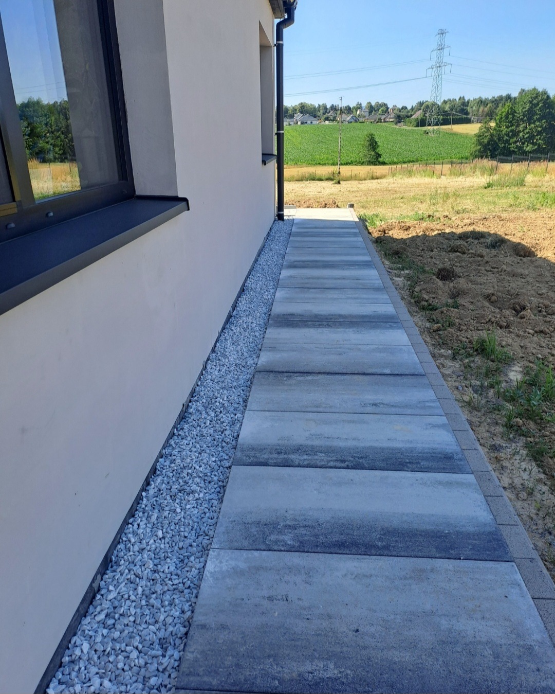 Nowoczesny chodnik z szarych płyt betonowych ułożony wzdłuż jasnej elewacji domu, z dekoracyjnym pasem białego żwiru. W tle zielone pole i błękitne niebo.
