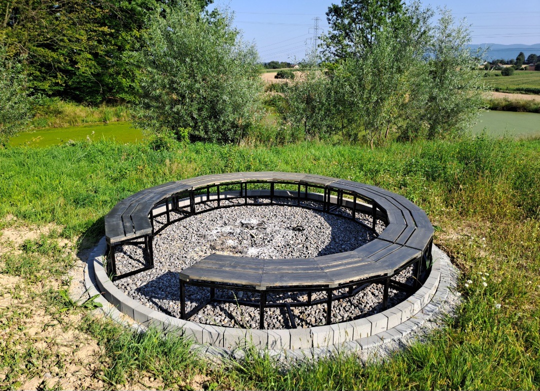 Ognisko otoczone ławkami z ciemnego drewna i kamieniami, na tle zieleni i stawu. Okrągła konstrukcja z siedziskami, idealna do spotkań na świeżym powietrzu.