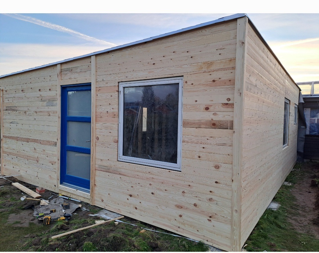 Drewniany budynek w trakcie budowy, z widocznymi deskami elewacyjnymi, niebieskimi drzwiami i białymi oknami. Na ziemi narzędzia budowlane.