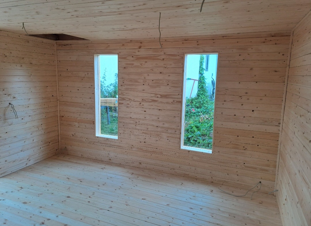 Wnętrze domu wykończone boazerią: ściany, podłoga i sufit z jasnego drewna. Dwa okna z widokiem na zieleń. Otwór w suficie. Surowy stan wykończenia.