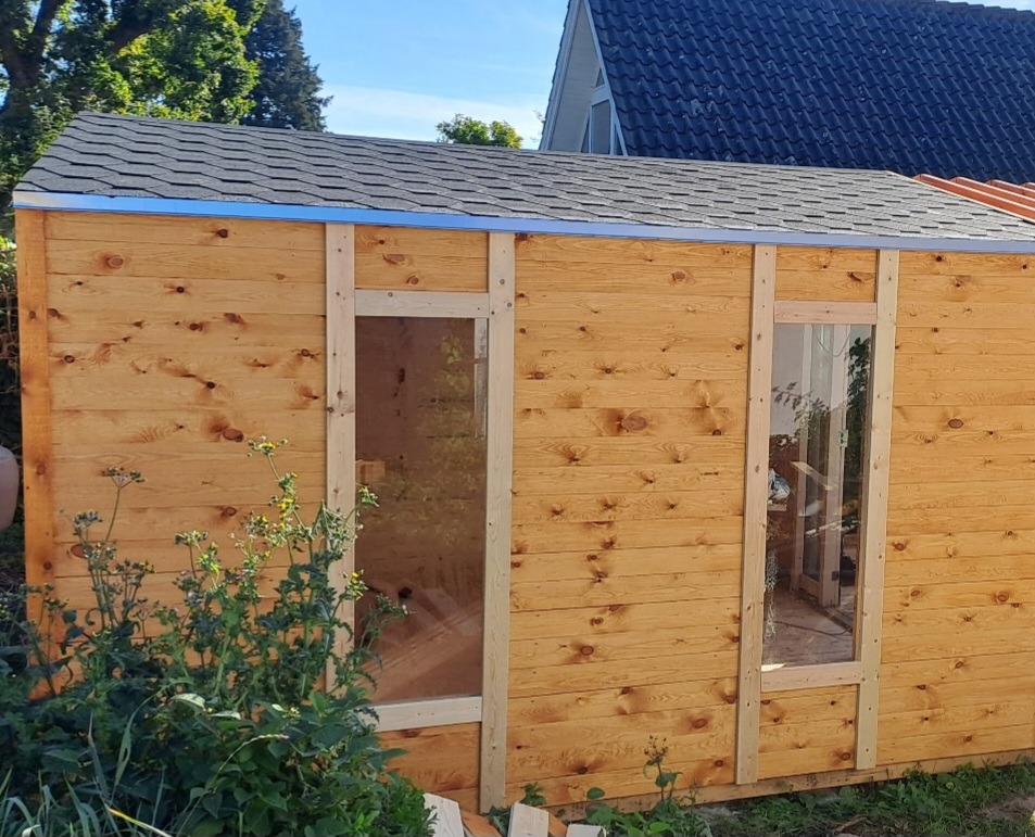 Drewniana konstrukcja z oknami i dachem z gontu bitumicznego, w trakcie budowy, widok z zewnątrz, jasne drewno, częściowo oszklone okna, niebieska listwa wykończeniowa.