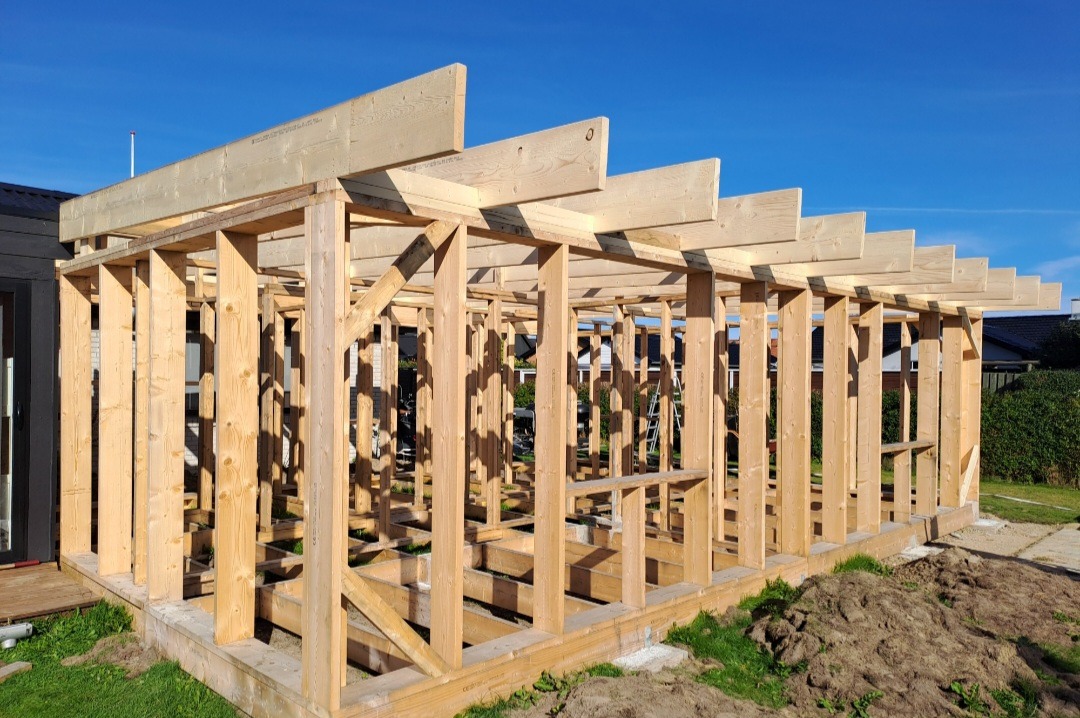 Drewniana konstrukcja szkieletowa budynku w trakcie budowy, widoczne słupy, belki i podwaliny, jasne drewno na tle błękitnego nieba.