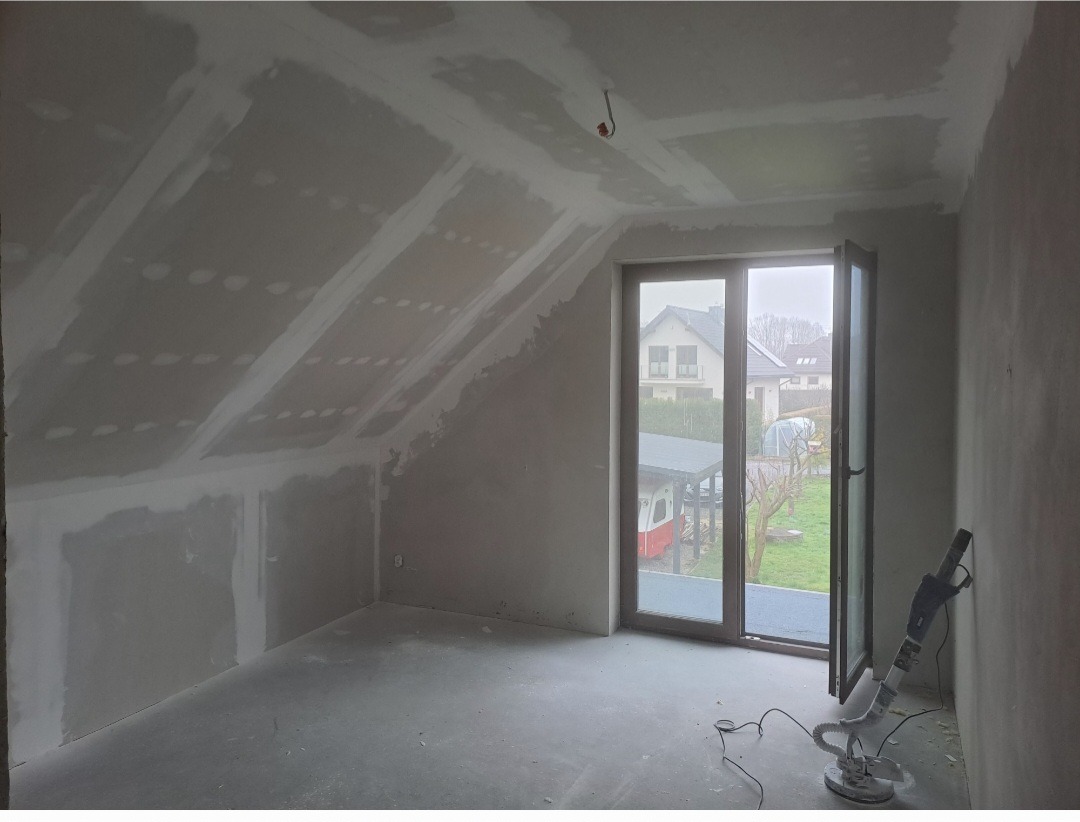 Poddasze po montażu płyt gipsowo-kartonowych i wstępnym szpachlowaniu, widoczna szlifierka do gipsu przy ścianie, okno balkonowe z widokiem na ogród.