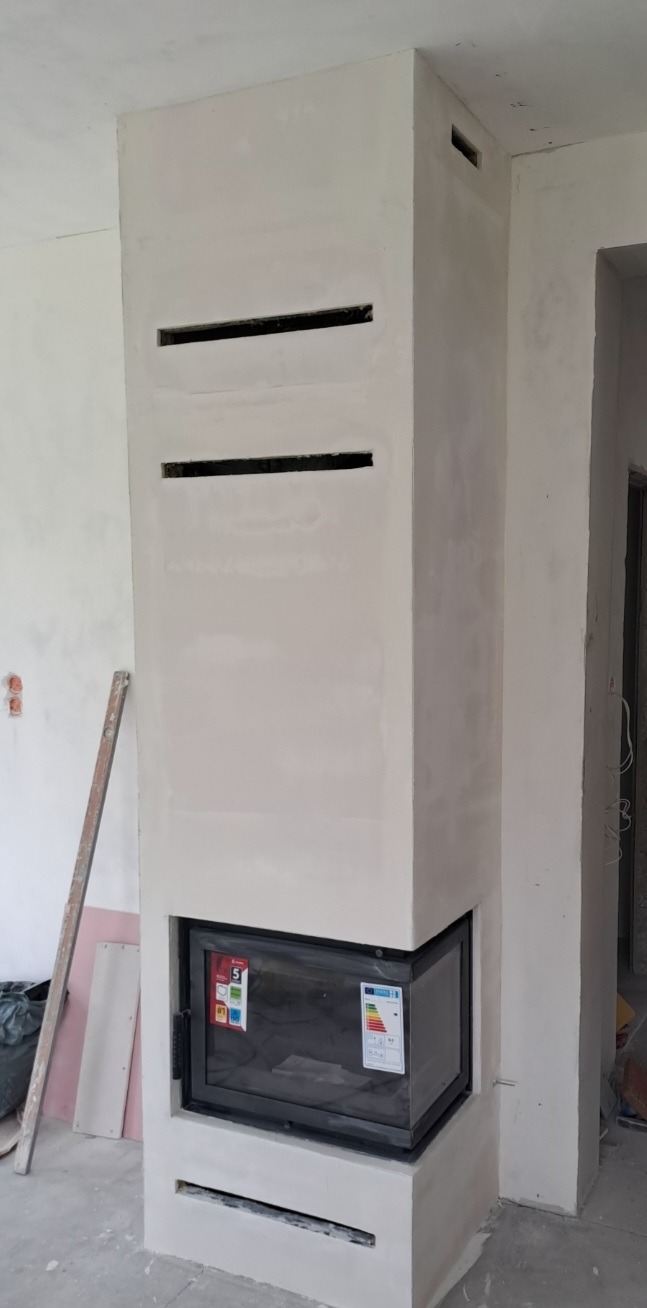 Obudowa kominka po szpachlowaniu, z widocznymi szczelinami wentylacyjnymi i szybą kominkową. Ściany w trakcie wykończenia, widoczne narzędzia.