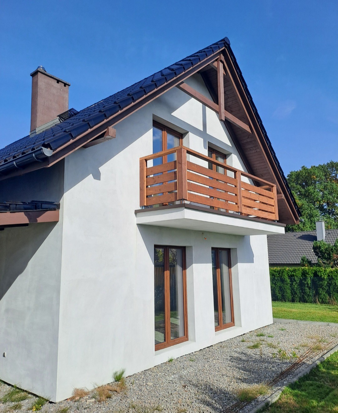 Biały dom z drewnianym balkonem i brązowymi oknami w słoneczny dzień. Widoczny fragment dachu z kominem i otoczenie domu.