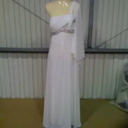 Manekin prezentuje długą, białą suknię wieczorową na jedno ramię z ozdobnym pasem z cekinów, sfotografowaną w magazynowym otoczeniu z blach falistych w tle.