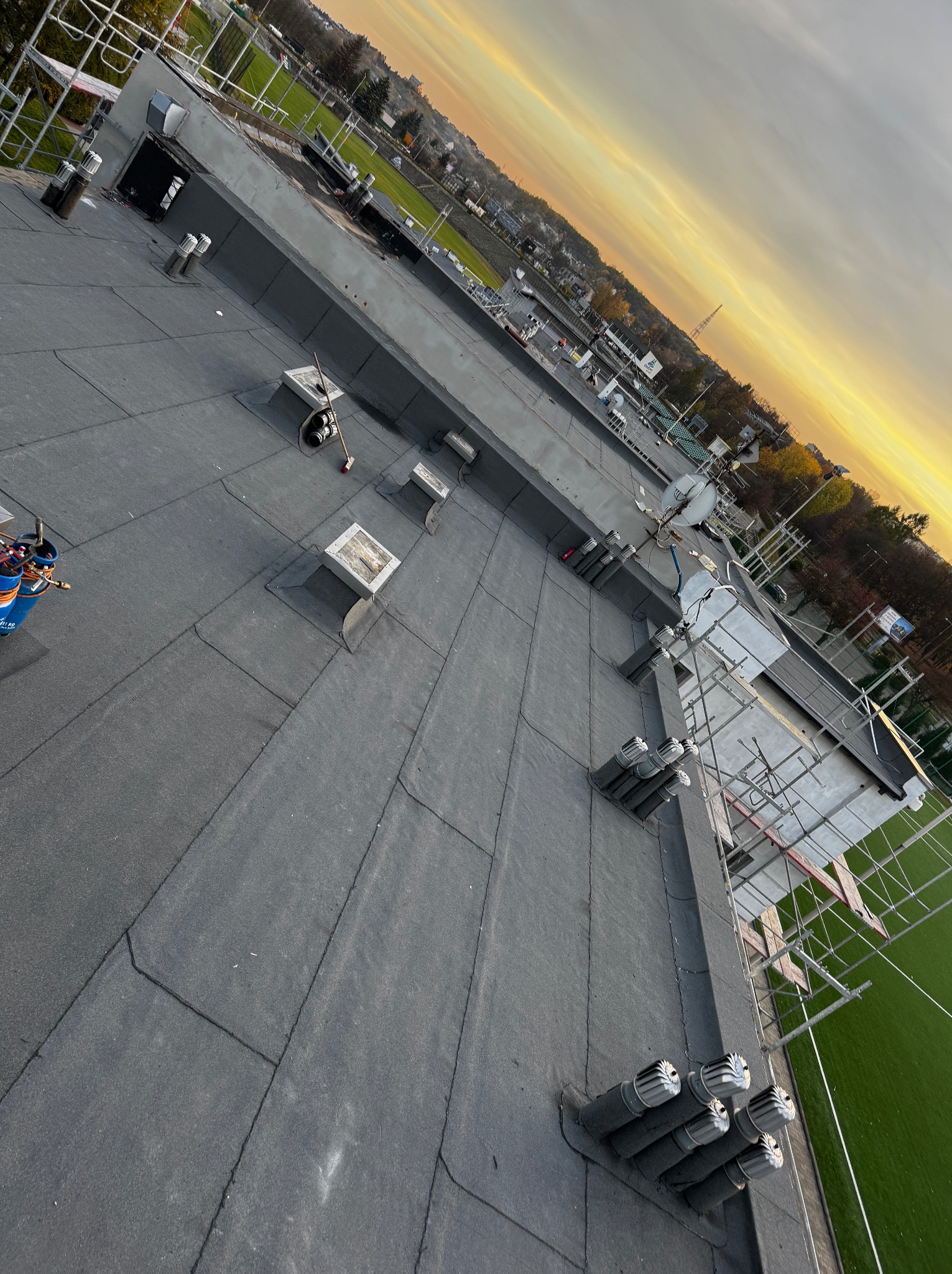 Płaski dach pokryty papą termozgrzewalną z kominkami wentylacyjnymi i narzędziami dekarskimi, widok z góry, w tle zachodzące słońce.