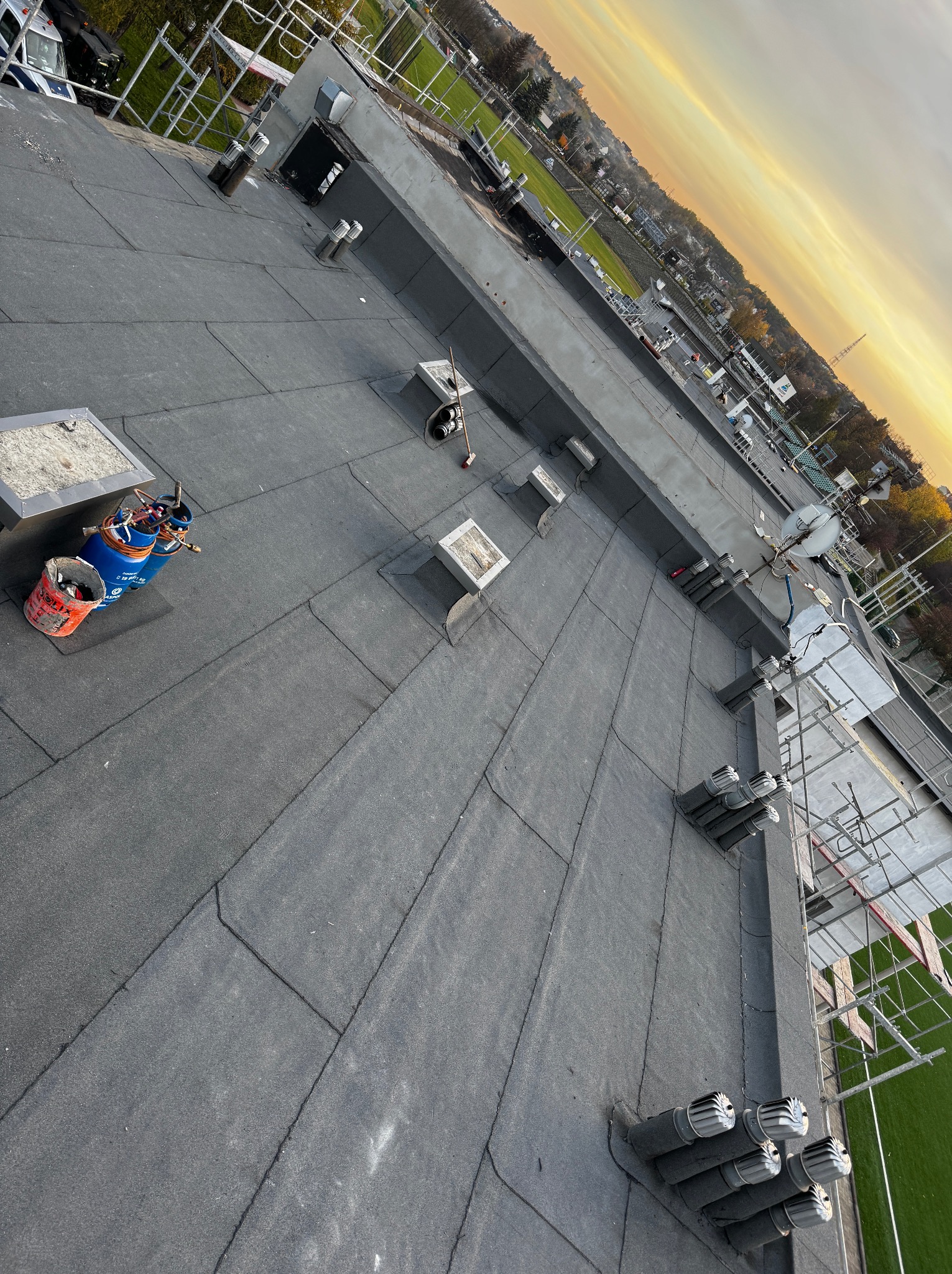 Widok z góry na płaski dach pokryty papą termozgrzewalną, z widocznymi kominkami wentylacyjnymi, butlami gazowymi i narzędziami dekarskimi na tle zachodzącego słońca.