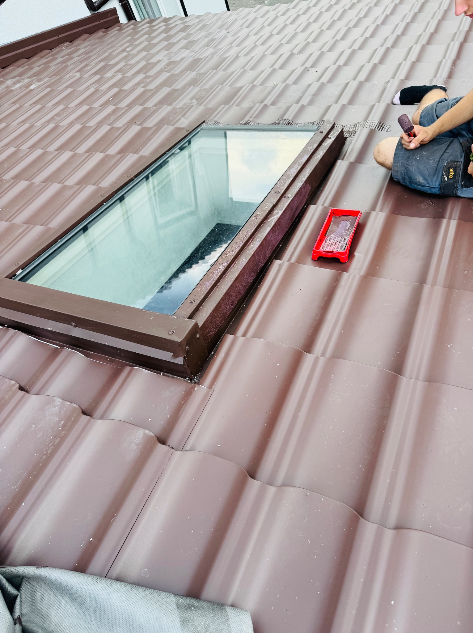 Malowanie blachodachówki wokół okna dachowego brązową farbą. Widoczny wałek i kuweta z farbą. Praca wykonywana na dachu.