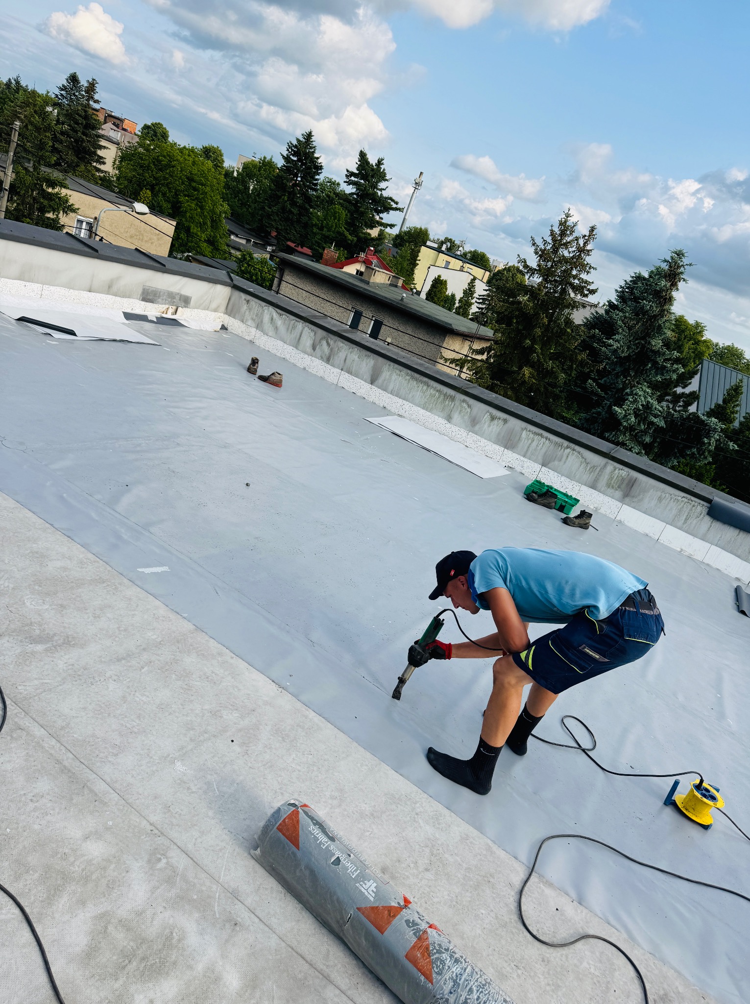 Pracownik w trakcie zgrzewania membrany na płaskim dachu, widoczne narzędzia i rolka materiału. W tle zabudowa miejska i zieleń.