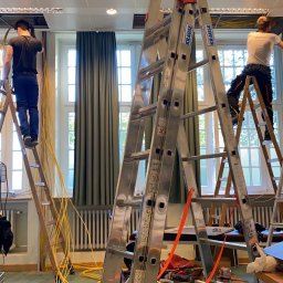 Damian Głąb - Dwóch elektryków na drabinach montuje okablowanie pod sufitem w jasnym pomieszczeniu z oknami. Widoczne zwisające kable i narzędzia. Ujęcie z dołu, podkreślające wysokość.