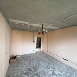 Damian Głąb - Pokój w trakcie remontu z widocznymi przewodami elektrycznymi na ścianach i suficie, surowy betonowy sufit, wisząca lampa. Widoczne gniazdka elektryczne.