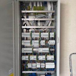 Damian Głąb - Otwarta szara rozdzielnica elektryczna EATON z rzędami zabezpieczeń, starannie ułożonymi kolorowymi przewodami i niebieskimi peszlami. Obok widoczna część metalowej drabiny.