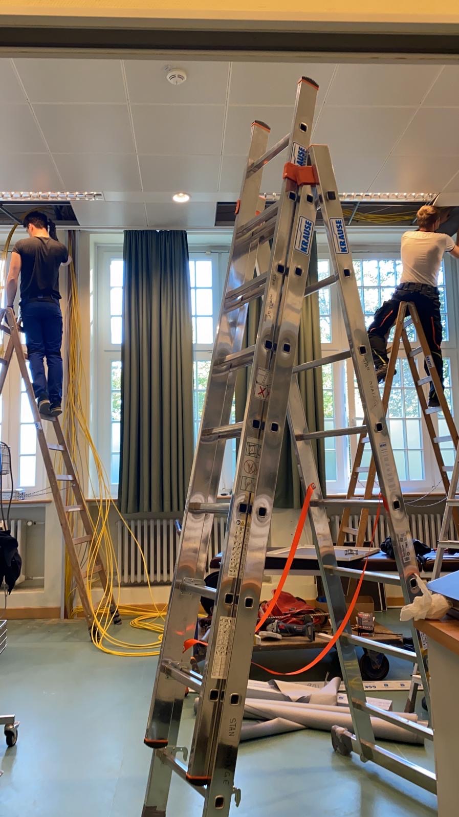 Dwóch elektryków na drabinach montuje okablowanie pod sufitem w jasnym pomieszczeniu z oknami. Widoczne zwisające kable i narzędzia. Ujęcie z dołu, podkreślające wysokość.