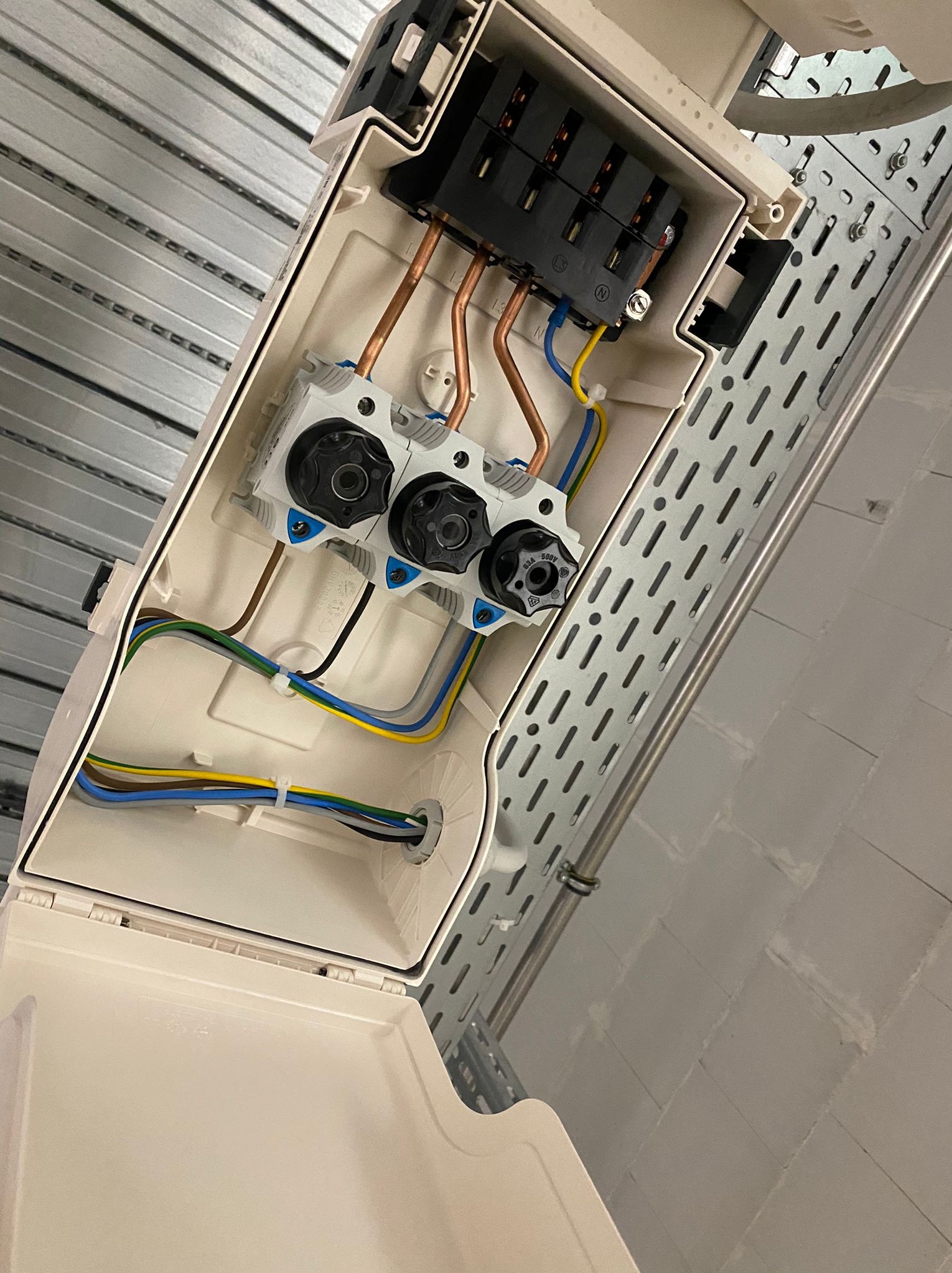 Otwarta, beżowa skrzynka elektryczna z bezpiecznikami i przewodami, zamontowana na metalowej konstrukcji. Widoczne kable w różnych kolorach i elementy instalacji.