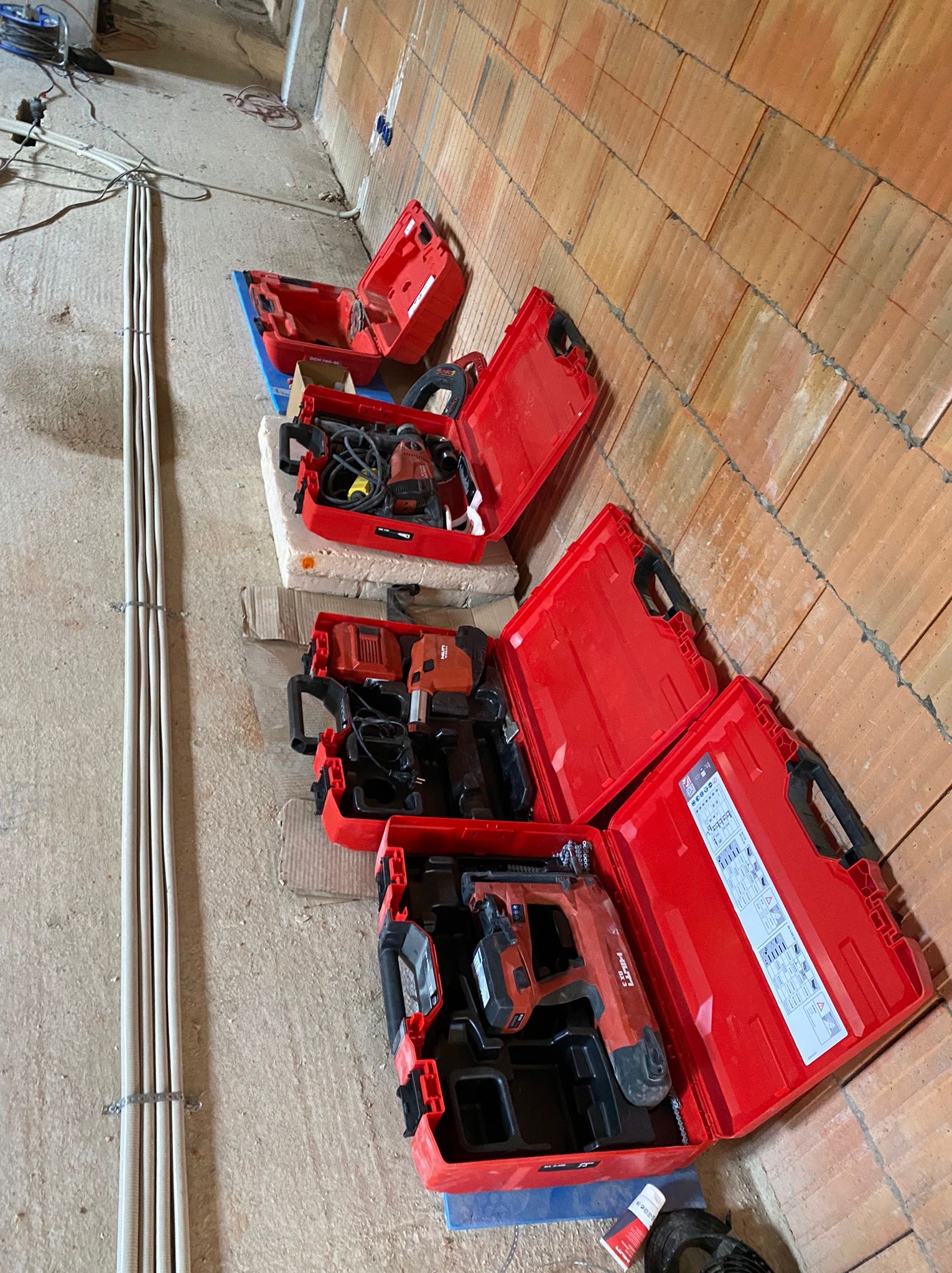 Rozłożone czerwone walizki z narzędziami Hilti na budowie, widoczne kable elektryczne przy ścianie z cegły. Przygotowanie do prac instalacyjnych.