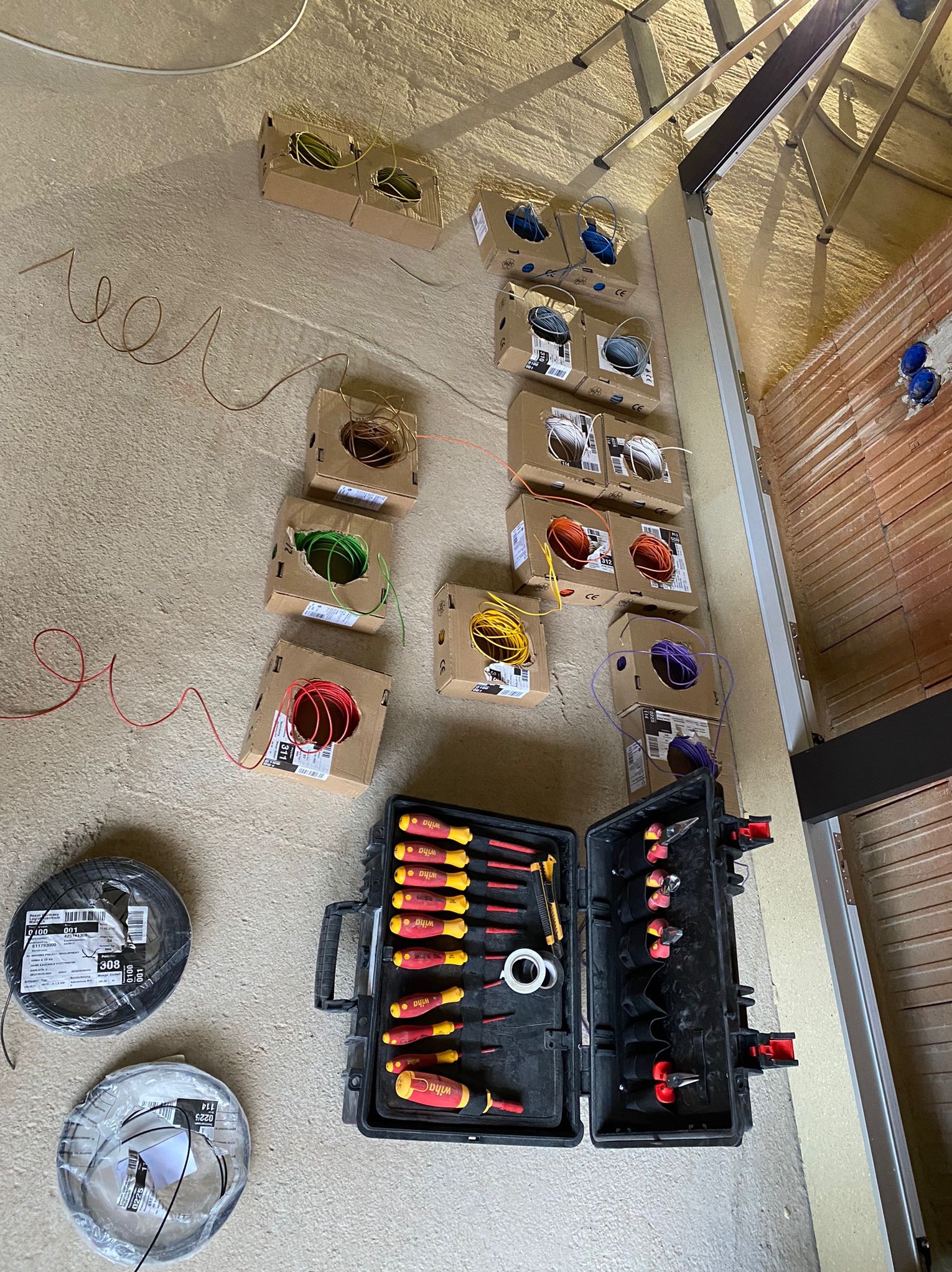 Rozłożone kable w kartonach, narzędzia Wiha i przewody na podłodze podczas prac elektrycznych w nowym budynku z widoczną konstrukcją dachu i ścianami z cegły.