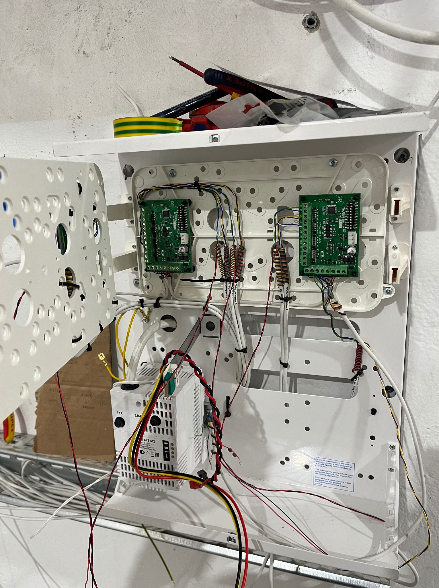 Otwarta skrzynka elektryczna z dwoma zielonymi modułami, licznymi przewodami i białym zasilaczem. Widoczne narzędzia i taśma izolacyjna na górze.