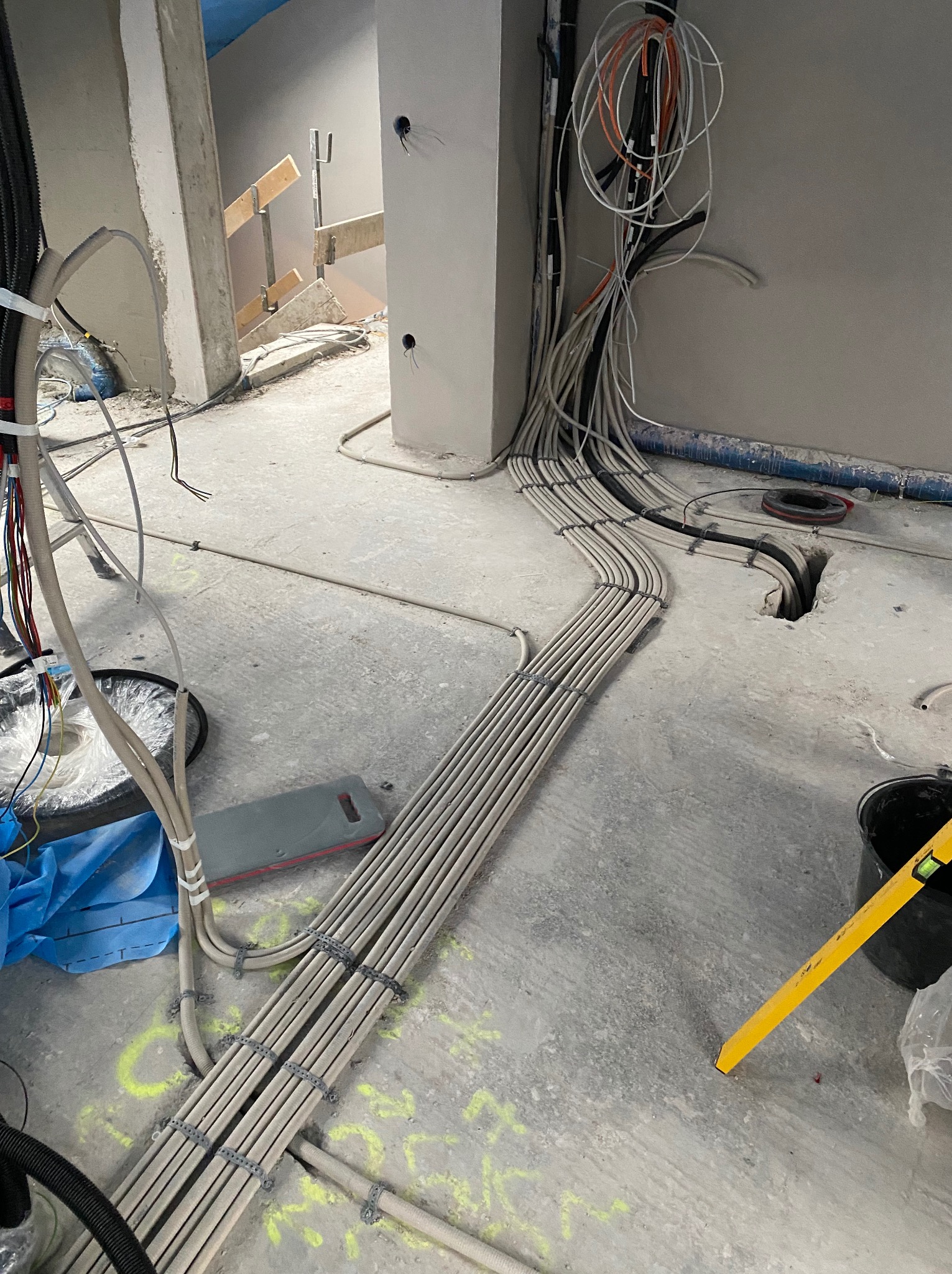 Szereg ułożonych kabli elektrycznych w peszlach na surowej podłodze, z widocznymi puszkami instalacyjnymi i poziomnicą, w trakcie prac instalacyjnych w budynku.