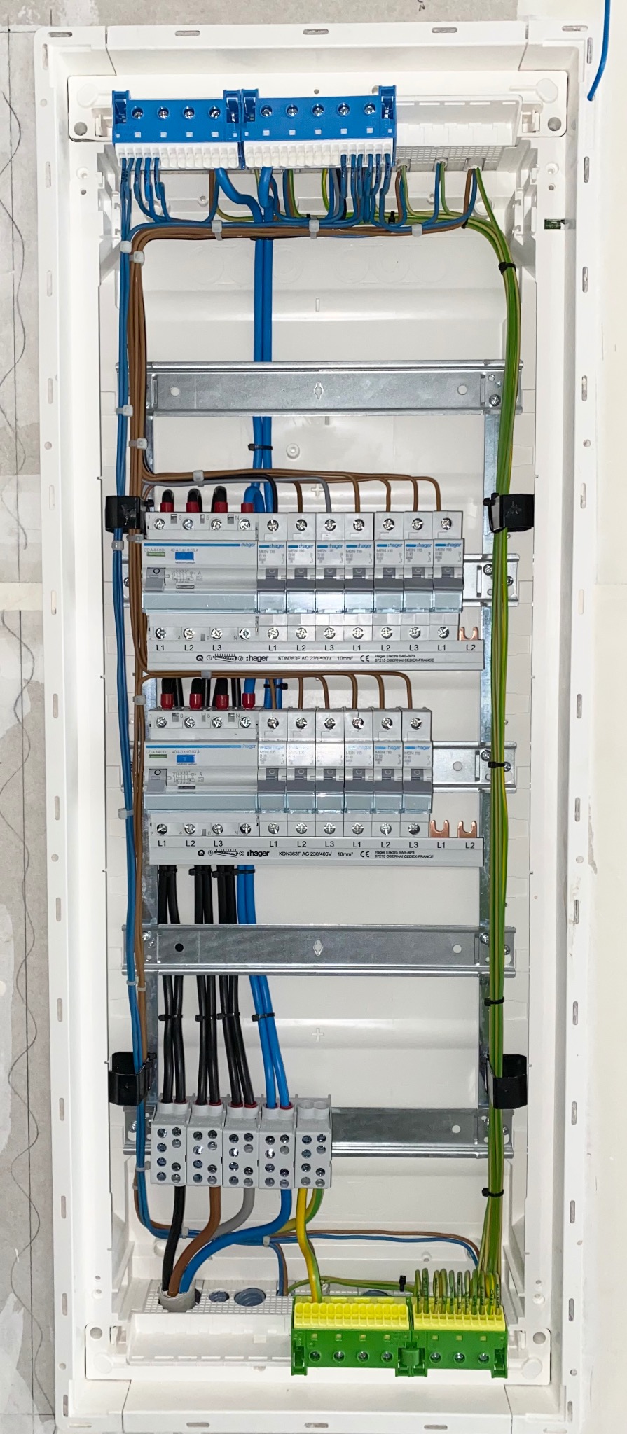 Rozdzielnica elektryczna Hager z zainstalowanymi bezpiecznikami i przewodami w kolorach: niebieskim, brązowym, czarnym i zielono-żółtym. Widoczne listwy zaciskowe.