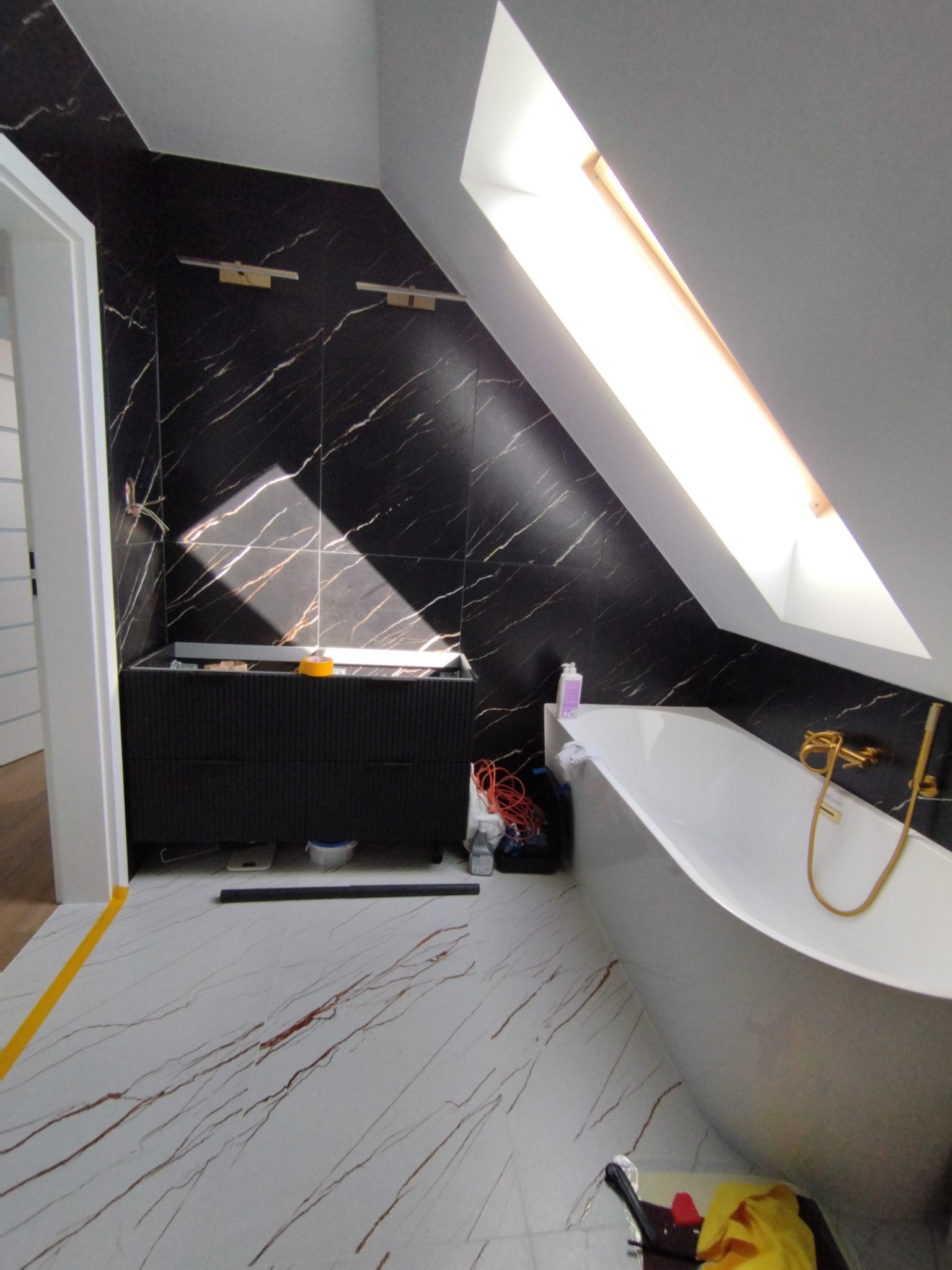Wykończona łazienka na poddaszu z czarnymi płytkami imitującymi marmur, białą wanną i podłogą z wzorem żył. Widoczne okno dachowe i złota armatura.